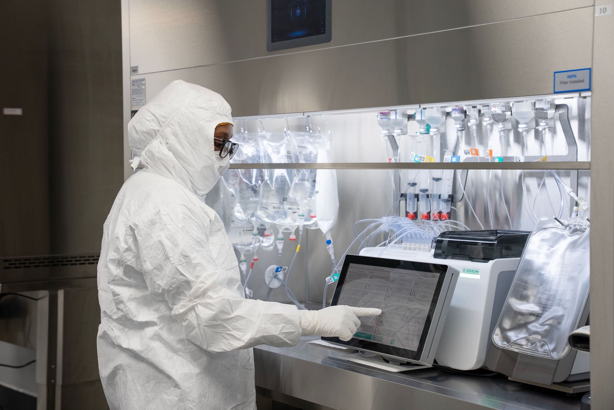 Cleanroom technician working at a touchscreen monitor in a sterile pharmaceutical compounding area with hanging IV bags and specialized equipment.