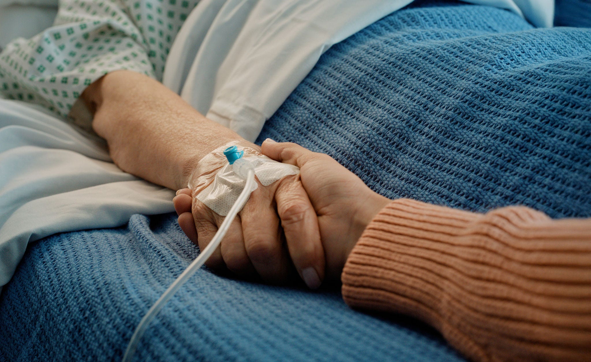 Patient in bed receiving IV therapy while another person holds the patient's hand.