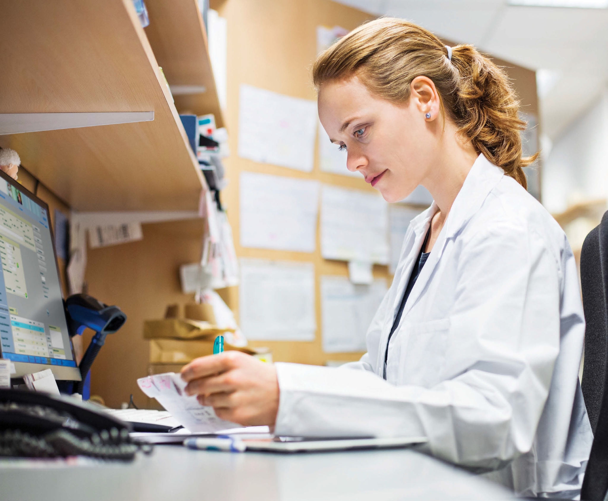 Healthcare professional seated at a workstation reviewing medication information while using a computer in a pharmacy or clinical setting.